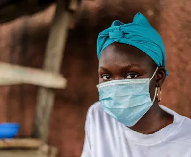 A portrait of a woman wearing a light blue surgical face mask. She is also wearing a turquoise head wrap and large gold hoop earrings.